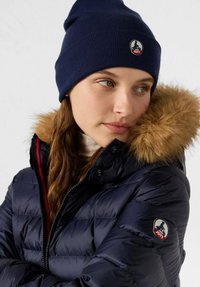 Giacca a vento imbottita blu navy con cappuccio foderato in pelliccia, dotata di chiusura con cerniera e logo ricamato sulla manica. Berretto a maglia blu navy sulla testa.