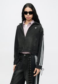 Μαύρο κοντό τζιν μπουφάν με φερμουάρ, λευκό λογότυπο και ριγωτές λεπτομέρειες στα μανίκια. Συνδυάζεται με μαύρο παντελόνι με διακοσμητικές τρύπες.