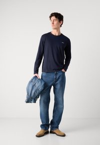 Navy long-sleeve shirt, blue denim jeans, beige shoes. Holding a denim jacket, slim fit, flat front, casual design, plain fabric.
