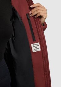 Chaqueta acolchada burdeos con interior negro, que cuenta con un bolsillo lateral con cremallera y una etiqueta que dice "Un cálido abrazo hecho de tela."