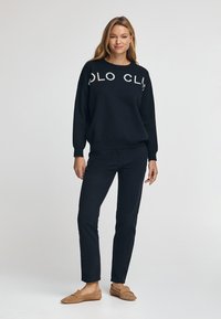 Svart sweatshirt med vita bokstäver "POLO CL" över framsidan, i kombination med mörka byxor och tanfärgade slip-on skor, stående framför en neutral bakgrund.