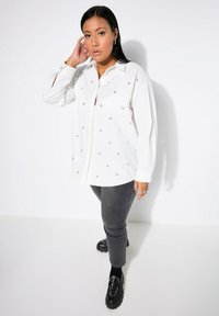 Camisa blanca de botón con tachuelas plateadas, de manga larga y corte relajado, combinada con jeans gris oscuro y zapatos de plataforma negros.