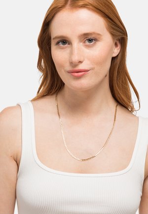 Collier en chaîne dorée avec un design plat et maillé reposant sur une clavicule nue. L'arrière-plan est uni pour mettre en valeur les détails du bijou.