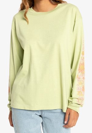 Vrouw draagt een lichtgroen shirt met lange mouwen met roze en gele bloemenpatronen op de mouwen en lichtblauwe spijkerbroek, gekort boven de taille.