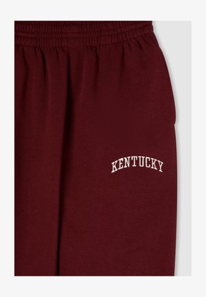 Pantaloni della tuta color bordeaux con cintura elastica, caratterizzati da testo ricamato bianco con la scritta "KENTUCKY" in un design ad arco. Tessuto morbido e testurizzato.