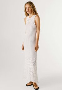 Žena stojaca, oblečená v bielych bezrukávových maxi pletených šatách s vzorovaným lemom a hnedej sandále na jednoduchom pozadí.