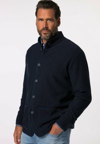 Marinblå stickad jacka med knappar, ståkrage och två framfickor. Tygets struktur är ribbad med kontrasterande kantdetaljer.