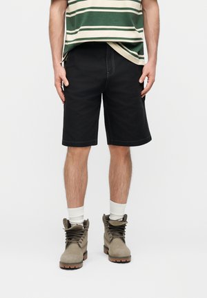 Férfi fekete térd alatti rövidnadrágban, fehér zokniban, bézs fűzős bakancsban és zöld–bézs csíkos ingben.