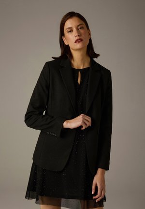 Schwarzer Blazer mit dezentem Nadelstreifenmuster, kombiniert mit einem kurzen schwarzen Kleid mit Schlüsselloch-Ausschnitt und gepunkteten Akzenten.