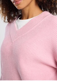 Maglione rosa lavorato a maglia con scollo a V, caratterizzato da una texture a coste e una vestibilità comoda, indossato sopra una maglietta bianca.