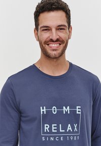 Granatowa koszula z długim rękawem z jasnoniebieskim nadrukiem tekstu "HOME RELAX SINCE 1989" w prostokątnym wzorze. Miękka tekstura materiału.