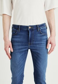 Μπλε τζιν παντελόνι με εφαρμοστή σχεδίαση, έχοντας πέντε τσέπες, φερμουάρ και κουμπί κλεισίματος με λογότυπο επωνυμίας.