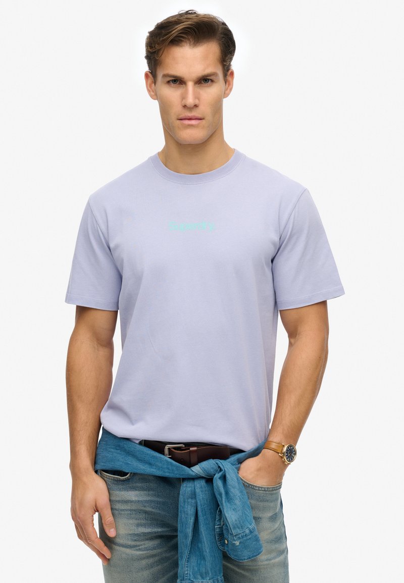 Camiseta de algodón morado claro con mangas cortas, que presenta el logo de "Superdry" en tono teal. Combinada con una camisa de denim atada a la cintura y jeans desgastados.