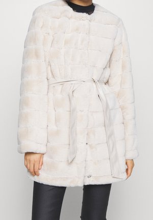 Persona che indossa un cappotto trapuntato in pelliccia sintetica beige chiaro con cintura in tessuto annodata in vita e pantaloni neri.