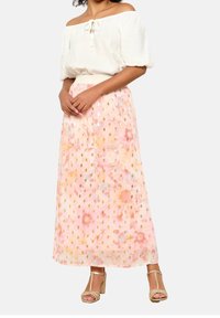 Blouse crème décolletée portée avec une longue jupe fluide rose pastel ornée de motifs floraux et d'accents dorés, accompagnée de talons dorés.