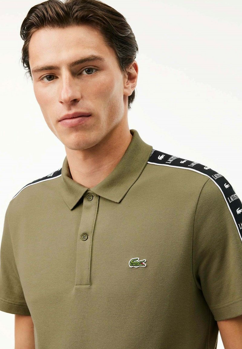 Polo vert olive avec un col classique, patte de boutonnage à trois boutons et une étiquette en tissu ; présente des rayures noires contrastantes sur les épaules.