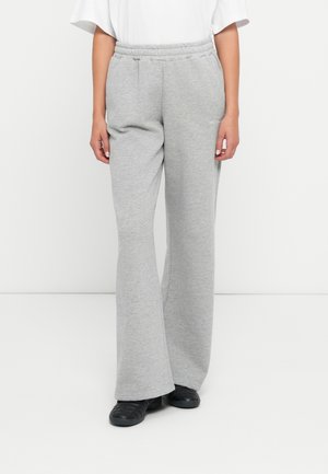 Personne portant un pantalon de jogging gris clair à jambes larges, des chaussures noires et un haut blanc sur un fond blanc uni.