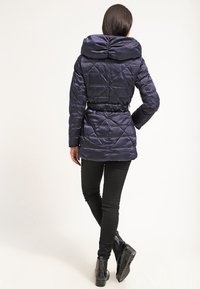 Marineblå puffjakke med quiltet mønster, stor hætte, elastisk talje og sorte detaljer. Båret over sorte skinny bukser og støvler.