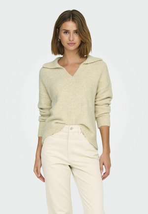 Lichtbeige trui met een v-hals en zachte kraag. Gebreide textuur en lange mouwen. Gecombineerd met beige broeken.