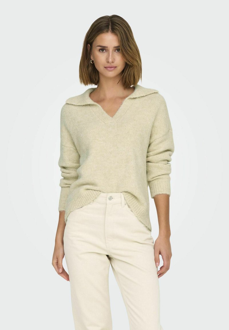 Lichtbeige trui met een v-hals en zachte kraag. Gebreide textuur en lange mouwen. Gecombineerd met beige broeken.