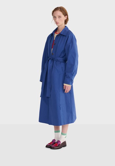 Femme debout portant un long manteau bleu ceinturé, des chaussettes blanches avec des rayures vertes et des mocassins rouge foncé à pompons sur fond blanc.