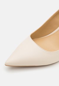Kärkikuvioinen korkokenkä pehmeästä beige nahasta, jossa on sileä pinta ja matala leikkuri. Ominaisuutena minimalistisella suunnittelulla ja tasaisella pohjalla.