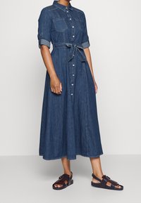 Woman wearing a long denim button-up dress with rolled sleeves and a tied waist belt, paired with dark open-toe sandals.