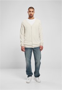 Beige gestrickte Strickjacke mit V-Ausschnitt, ausgestattet mit einer Knopfleiste, langen Ärmeln und einer strukturierten Oberfläche, kombiniert mit hellblauen Jeans und weißen Sneakers.