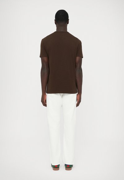 Barna rövid ujjú pamutpóló, fehér egyenes szárú nadrággal és színes slip-on cipővel párosítva, hátulnézetből.