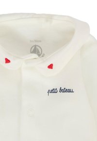 Kremowa bawełniana koszulka polo z złożonym kołnierzykiem, ozdobiona haftem w czerwone serce i granatowym napisem "petit bateau". Guziki są białe.