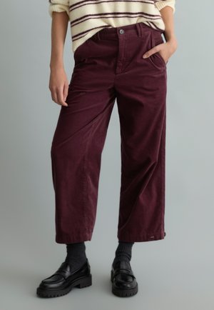 Personne portant un pantalon large bordeaux, des chaussettes noires, des mocassins noirs épais et un pull crème avec des rayures noires. Une main dans la poche.