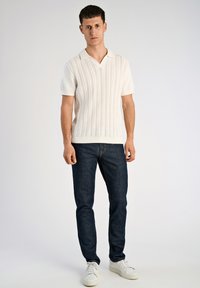 Λευκό πλεκτό πόλο με κοντό μανίκι, V-λαιμόκοψη και ραβδωτή υφή, συνδυασμένο με σκούρο μπλε τζιν στενής γραμμής και λευκά αθλητικά παπούτσια.