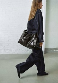 Vrouw die binnen loopt, gekleed in een donkere spijkeroutfit en zwarte schoenen, met een grote zwarte leren tas over haar schouder.
