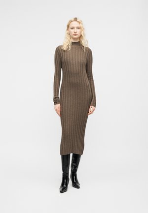 Šviesiaplaukė moteris dėvi rudą, dryžuotą, megztą ilgomis rankovėmis midi ilgio suknelę ir juodus aukštus iki kelių batus, stovėdama priešais paprastą baltą foną.