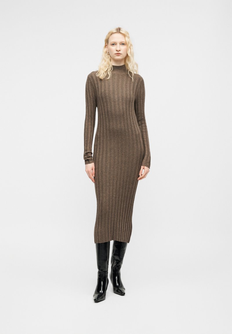 Femme blonde portant une robe midi marron à manches longues en maille côtelée et des bottes noires montantes, debout devant un fond blanc uni.