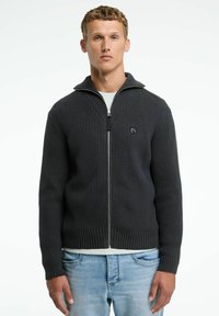 Dunkelgrauer Reißverschluss-Pullover mit geripptem Muster, hohem Kragen und einem Emblem auf der linken Brust. Über einem hellen Hemd getragen und mit blauen Jeans kombiniert.
