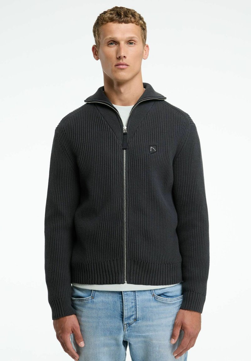 Dunkelgrauer Reißverschluss-Pullover mit geripptem Muster, hohem Kragen und einem Emblem auf der linken Brust. Über einem hellen Hemd getragen und mit blauen Jeans kombiniert.
