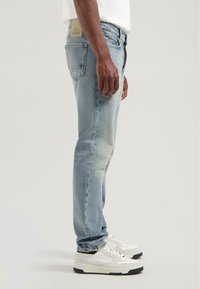 Man in lichte blauwe jeans en witte sneakers, staande opzij tegen een effen lichte achtergrond.