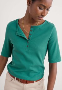 Zeleni rebrasti T-shirt s okruglim izrezom, bočnim gumbima i kratkim rukavima. Stiliziran s smešnim pojasom koji ima suptilne teksturne detalje.