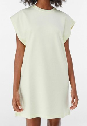 Mujer que lleva un vestido suelto, sin mangas y amarillo pálido con hombreras, de pie frente a un fondo blanco liso.