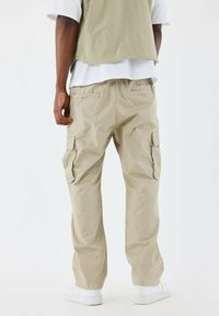 Pantalones cargo de color beige claro con bolsillos laterales, diseño de pierna ajustada, hechos de una tela texturizada, llevados con un chaleco verde oliva suelto.