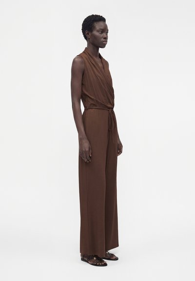 Mannequin mince portant une combinaison marron sans manches à jambes larges avec des sandales plates noires, debout sur fond blanc.