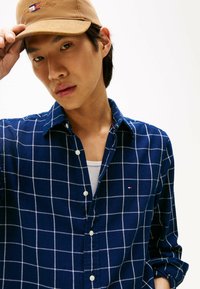 Joven con una gorra marrón y una camisa de cuadros azules sobre una camiseta blanca, ajustándose la gorra con una mano.