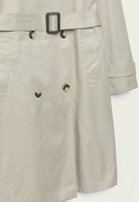 Trench-coat beige à texture lisse, à double boutonnage, avec des boutons noirs et une ceinture assortie dotée d'une boucle rectangulaire.