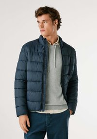 Mornarska puhovka z izšitjem, visokim ovratnikom in sprednjim zadrgo. Oblikovana čez sivo majico s kapuco in bež srajco spodaj.