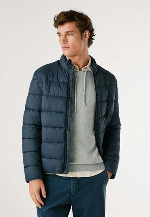 Marineblauwe pufferjack met een gequilte stof, hoge kraag en een ritssluiting aan de voorkant. Gelaagd over een grijze sweatshirt met trekkoord en een beige shirt eronder.