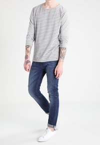 Camisa de manga larga a rayas en gris y blanco, de tejido suave, con cuello redondo. Combinada con jeans azules oscuros y zapatillas blancas. Accesorios mínimos.