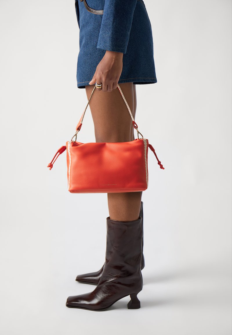 Bolso de piel roja con superficie texturada, que presenta un ribete trenzado y borlas. Combinado con botas cortas marrones y un vestido mini de mezclilla.