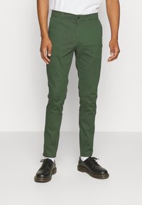 Chinos verdi realizzati in tessuto misto cotone con vestibilità slim. Presentano una chiusura a bottone, tasche laterali e una texture liscia. Abbinati a scarpe nere.