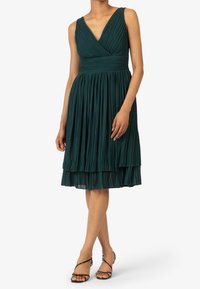 Teal plisserad klänning med djup V-ringning, figurnära överdel och lager på lager kjol. Tillverkad av textil med struktur, med ett brett midjeband.